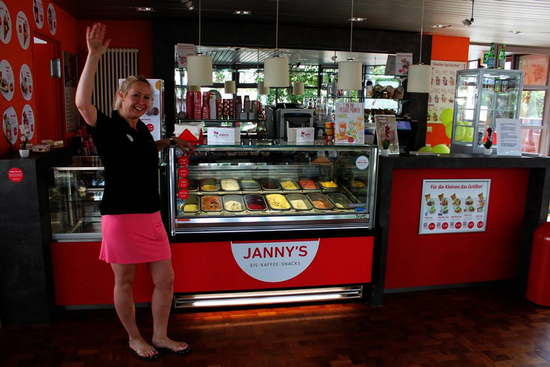 Eine Frau steht in einem Eiscafé vor einer Eistheke, auf der "Janny's Eis – Kaffee – Snacks" steht. Sie trägt ein schwarzes Oberteil und einen pinken Rock und hebt eine Hand zum Gruß. In der Theke sind verschiedene Sorten Eis zu sehen. Im Hintergrund befinden sich Werbeschilder und eine moderne Innenausstattung in Rot und Dunkelgrau.