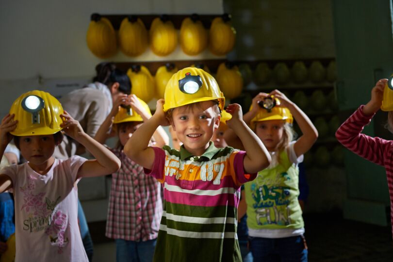 Das Bild zeigt eine Gruppe von Kindern, die gelbe Helme mit integrierten Grubenlampen tragen. Sie stehen in einem Raum mit vielen Helmen an der Wand im Hintergrund. Ein Kind im Vordergrund lächelt und setzt seinen Helm auf, während andere Kinder ebenfalls ihre Helme anpassen. Die Stimmung wirkt fröhlich und aufgeregt.