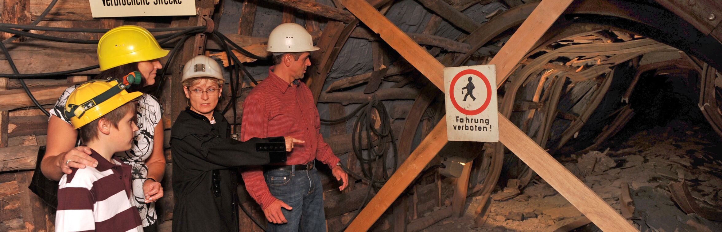 Das Bild zeigt eine Frau im schwarzen Hemd, die eine Familie durch einen alten Stollen im Anschauungsbergwerk Oelsnitz führt. Die Familie trägt Helme und steht vor einer gesperrten Strecke, die mit einem Schild „Fahrung verboten!“ markiert ist. Die Frau zeigt auf die verbrochene Strecke und erklärt vermutlich etwas über den Stollen. Die Wände des Stollens bestehen aus Holz, und die Beleuchtung sorgt für eine authentische, gedämpfte Bergwerksatmosphäre.