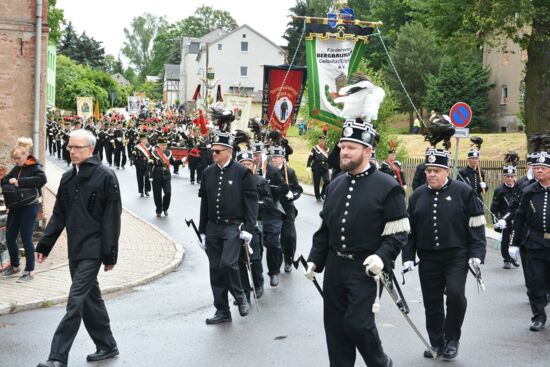 Ein weiterer Teil des Umzugs zeigt mehrere Bergleute in schwarzer Uniform, die eine große Fahne tragen, auf der „Förderverein Bergbaumuseum Oelsnitz/Erzgebirge e.V.“ steht. Hinter ihnen marschiert eine große Gruppe von Menschen, die ebenfalls in Uniformen gekleidet sind. Der Umzug findet in einer Wohngegend statt, mit Häusern und Bäumen im Hintergrund. Einige Zuschauer stehen am Straßenrand und beobachten das Geschehen.