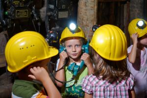 Das Bild zeigt mehrere Kinder, die gelbe Grubenhelme mit Lampen tragen. Ein Mädchen im Vordergrund hält ein altes Telefon mit beiden Händen an die Ohren. Die Kinder stehen in einem Bereich mit alten Maschinen und Schaltern, die an der Wand befestigt sind. Sie wirken konzentriert und neugierig, während sie die technischen Geräte erkunden.