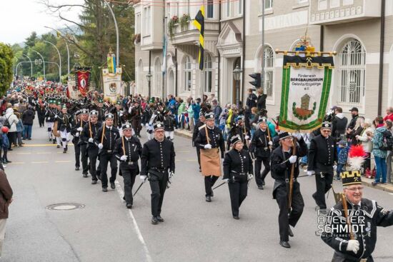 Ein Straßenumzug, bei dem Bergleute in schwarzen Uniformen und mit traditionellen Bergmannsmützen durch eine Stadt marschieren. Sie tragen Werkzeuge und Fahnen, während zahlreiche Zuschauer den Umzug von den Gehwegen aus beobachten. Im Hintergrund sind historische Gebäude und Laternenpfähle zu sehen. Eine große Fahne mit dem Wappen des Bergbaumuseums Oelsnitz ist gut sichtbar.