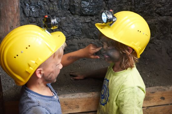 Das Bild zeigt zwei Kinder in einem Anschauungsbergwerk. Beide tragen gelbe Helme mit Grubenlampen, während eines der Kinder dem anderen vorsichtig Kohlenstaub ins Gesicht malt. Ihre Gesichter und Kleidung sind bereits von Kohlenstaub bedeckt, und sie scheinen sich dabei zu amüsieren. Im Hintergrund ist eine dunkle Wand aus Kohle zu sehen, die den Eindruck eines echten Bergwerks verstärkt.