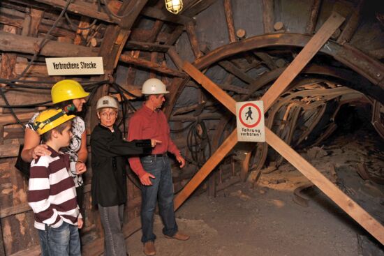 Das Bild zeigt eine Frau im schwarzen Hemd, die eine Familie durch einen alten Stollen im Anschauungsbergwerk Oelsnitz führt. Die Familie trägt Helme und steht vor einer gesperrten Strecke, die mit einem Schild „Fahrung verboten!“ markiert ist. Die Frau zeigt auf die verbrochene Strecke und erklärt vermutlich etwas über den Stollen. Die Wände des Stollens bestehen aus Holz, und die Beleuchtung sorgt für eine authentische, gedämpfte Bergwerksatmosphäre.