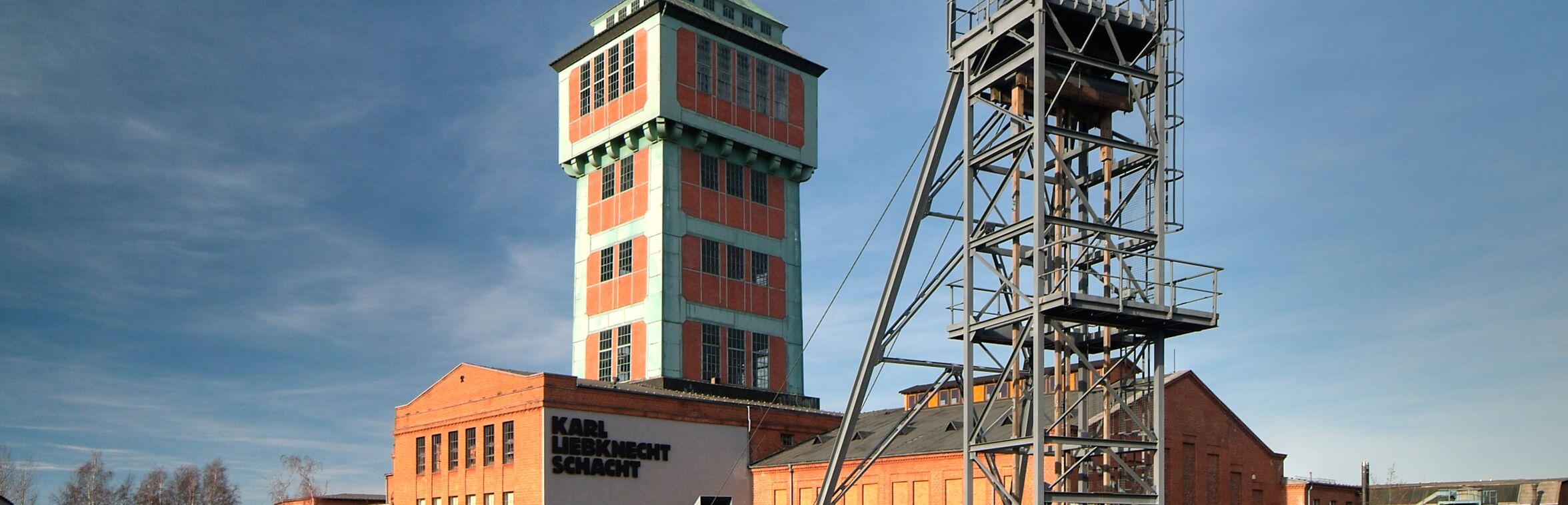 Das Bild zeigt eine Außenansicht des Bergbaumuseums Oelsnitz mit dem markanten Karl-Liebknecht-Schacht und einem hohen Förderturm. Im Vordergrund steht eine schwarze Dampflokomotive auf Schienen, umgeben von einem gepflasterten Platz. Der Himmel ist klar, und die Sonne scheint auf die roten Ziegelgebäude. Es gibt vereinzelt Büsche und Bäume, die die Umgebung beleben.