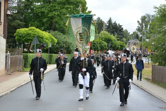 Zu sehen ist ein festlicher Umzug auf einer Straße, angeführt von Männern in traditioneller Bergmannskleidung, die schwarze Uniformen tragen. Einer der Männer in der Mitte trägt eine große grün-weiße Fahne mit einem Wappen. Im Hintergrund folgt eine größere Gruppe ebenfalls in Uniformen, und die Straße ist von grünen Bäumen gesäumt. Einige Zuschauer stehen am Straßenrand und beobachten den Umzug.