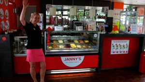 Eine Frau steht in einem Eiscafé vor einer Eistheke, auf der "Janny's Eis – Kaffee – Snacks" steht. Sie trägt ein schwarzes Oberteil und einen pinken Rock und hebt eine Hand zum Gruß. In der Theke sind verschiedene Sorten Eis zu sehen. Im Hintergrund befinden sich Werbeschilder und eine moderne Innenausstattung in Rot und Dunkelgrau.